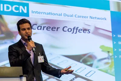 A speaker presents with microphone and clicker before a large Career Coffees screen, captured with crisp corporate photography and confident, open gestures.