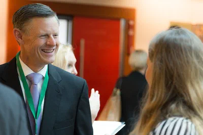 A smiling man in formal attire chats with another attendee during a networking break, warmly framed against red doors and a softly blurred crowd.