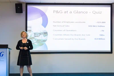 A presenter stands before a large corporate quiz slide, her poised stance and clear stage composition emphasizing polished conference photography by Alex.