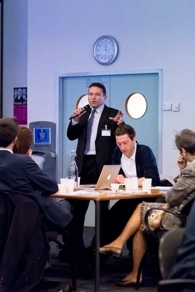 A standing participant speaks into a handheld microphone during a round-table session as colleagues listen, work on a laptop, and review materials.