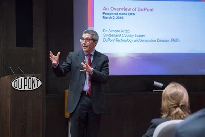 A business speaker gestures mid-presentation before a projected overview slide, captured in focused corporate event photography with audience members in the foreground.