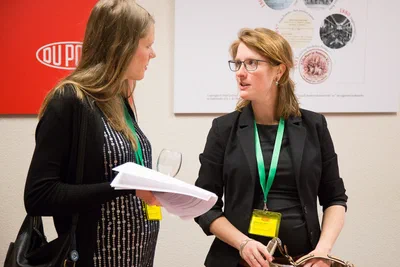 Two conference attendees wearing green lanyards exchange notes and conversation beside branded displays, creating a candid, professional networking portrait.