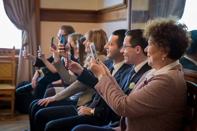 A seated row of guests raises phones and compact cameras in unison, smiling as they photograph an unseen moment in a warmly lit hall.