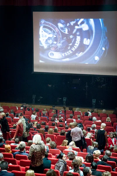 A bustling theatre audience settles into vivid red seats beneath a large projected watch image, captured in expansive event photography by Alex.