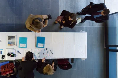 An overhead composition shows participants consulting documents around a white conference table, with colorful charts and clean geometric lines organizing the scene.