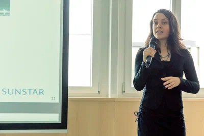 A poised speaker addresses the room with a handheld microphone beside a projected slide, captured in bright, natural conference photography.