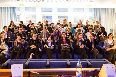 A lively conference audience settles into blue auditorium seats, with candid conversations and attentive expressions conveying the energy before a session.