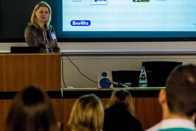 A presenter speaks from a wooden lectern beneath a large projection, framed through the audience for an immersive conference perspective.