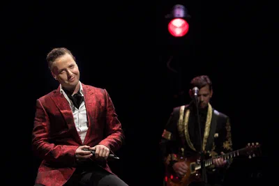 Under dramatic stage lighting, a vocalist in a red jacket smiles beside a guitarist, capturing the polished atmosphere of a live performance.