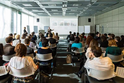 A wide conference view shows a full audience focused on a presenter and projected business slide in a bright, contemporary meeting room.