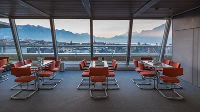 An empty meeting lounge with coral chairs overlooks rooftops, lake, and mountains, documenting a serene corporate venue before guests arrive.