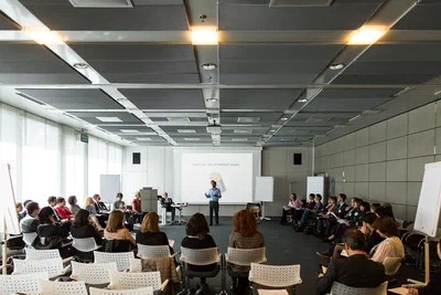 Participants form a broad circle around a central workshop leader, photographed by Alex in Geneva, Switzerland, in a bright modern conference room.