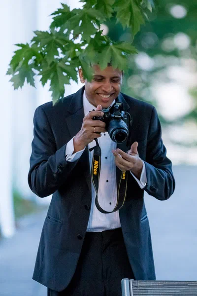 A smiling guest reviews a photograph on his camera beneath leafy branches, adding a playful behind-the-scenes moment to the event story.