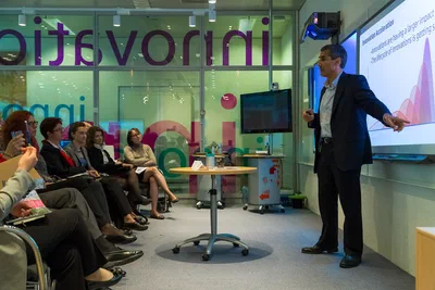A business speaker points toward a colorful presentation while a seated audience follows closely in an intimate, glass-walled conference space.