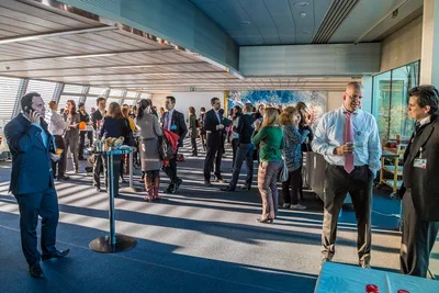 Professionals mingle in small groups at a sunlit networking reception, with long shadows and candid exchanges giving the corporate photography a dynamic feel.