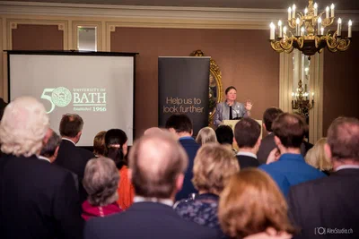 A speaker addresses a standing audience in an ornate chandelier-lit room, captured from behind the crowd for a strong sense of scale.