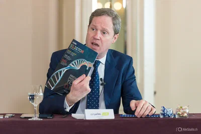 A seated panelist raises a magazine while speaking at a formal table, photographed by Alex in Geneva, Switzerland, with clean editorial clarity.