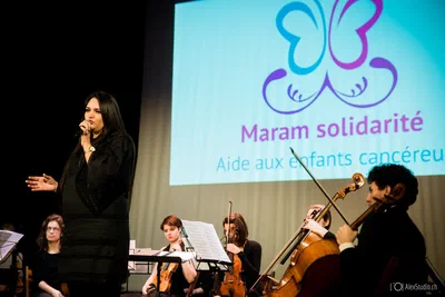 A vocalist performs beside a seated string ensemble as colorful event branding fills the screen, blending expressive stage presence with musical detail.