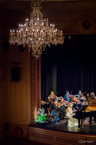 A pianist in a flowing gown performs with a chamber orchestra beneath a grand crystal chandelier in an elegant, atmospheric concert photograph.