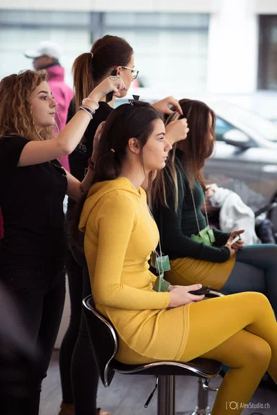 Stylists prepare seated participants' hair in a bright backstage setting, documenting the focused activity and careful details behind a live event.