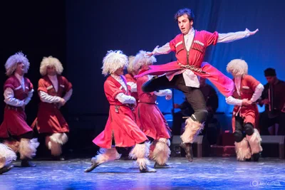 A dancer leaps at center stage while an ensemble performs in vivid traditional costumes, frozen sharply against rich blue theatrical lighting.