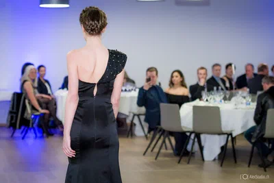 A model presents a black one-shoulder gown while seated guests watch from round tables, captured in polished event photography by Alex.