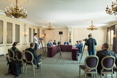 Camera crews document a formal panel discussion in an elegant chandeliered room, revealing both the speakers and the production behind the conference.