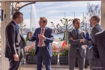 Four suited attendees hold two animated conversations on a sunlit terrace, with water and sculptural trees creating a refined corporate setting.