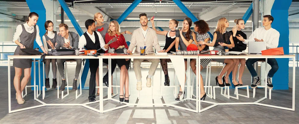 A lively corporate team gathers around long white tables in a bright modern office with bold blue structural beams.