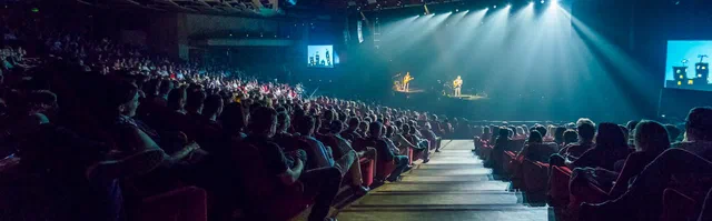 A packed auditorium watches two performers on a distant stage, illuminated by sweeping blue-white spotlights during a large Geneva event.