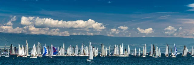 A panoramic fleet of sailboats stretches across Geneva's blue lake, framed by distant mountains and billowing white clouds.