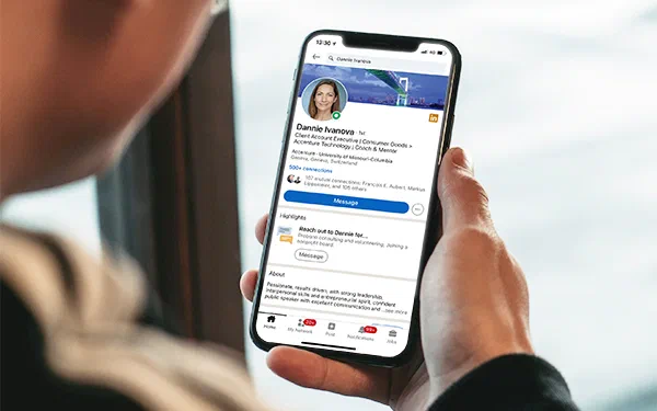 A smartphone displays a LinkedIn profile with a professional headshot while the user holds the device near a bright window.