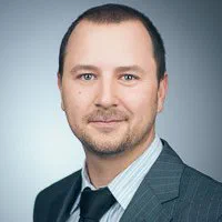 A man with short dark hair and light stubble wears a charcoal suit and black tie for a composed corporate portrait.