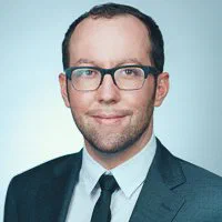 A man wearing rectangular glasses, a charcoal suit, and black tie smiles subtly in a crisp LinkedIn photo on light gray.