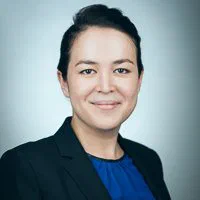 A woman smiles gently in a black blazer and vivid blue top for a polished LinkedIn photo against a cool gray backdrop.