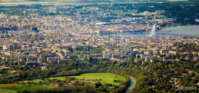 A wide elevated view surveys Geneva, its lake and dense cityscape beneath layered blue mountains and a luminous, cloud-filled sky.